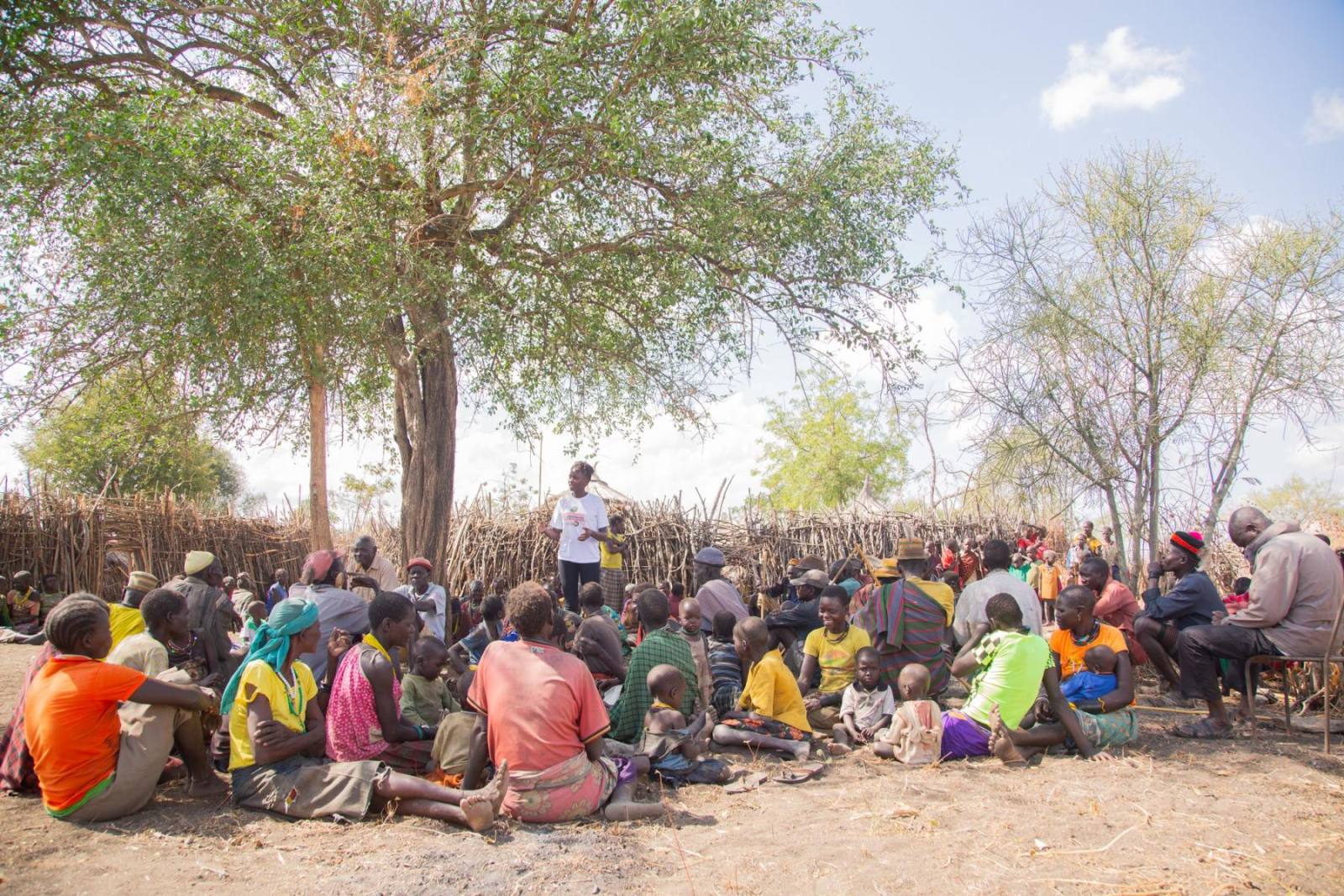 A large community group seated in the shade of a tree for a dialogue